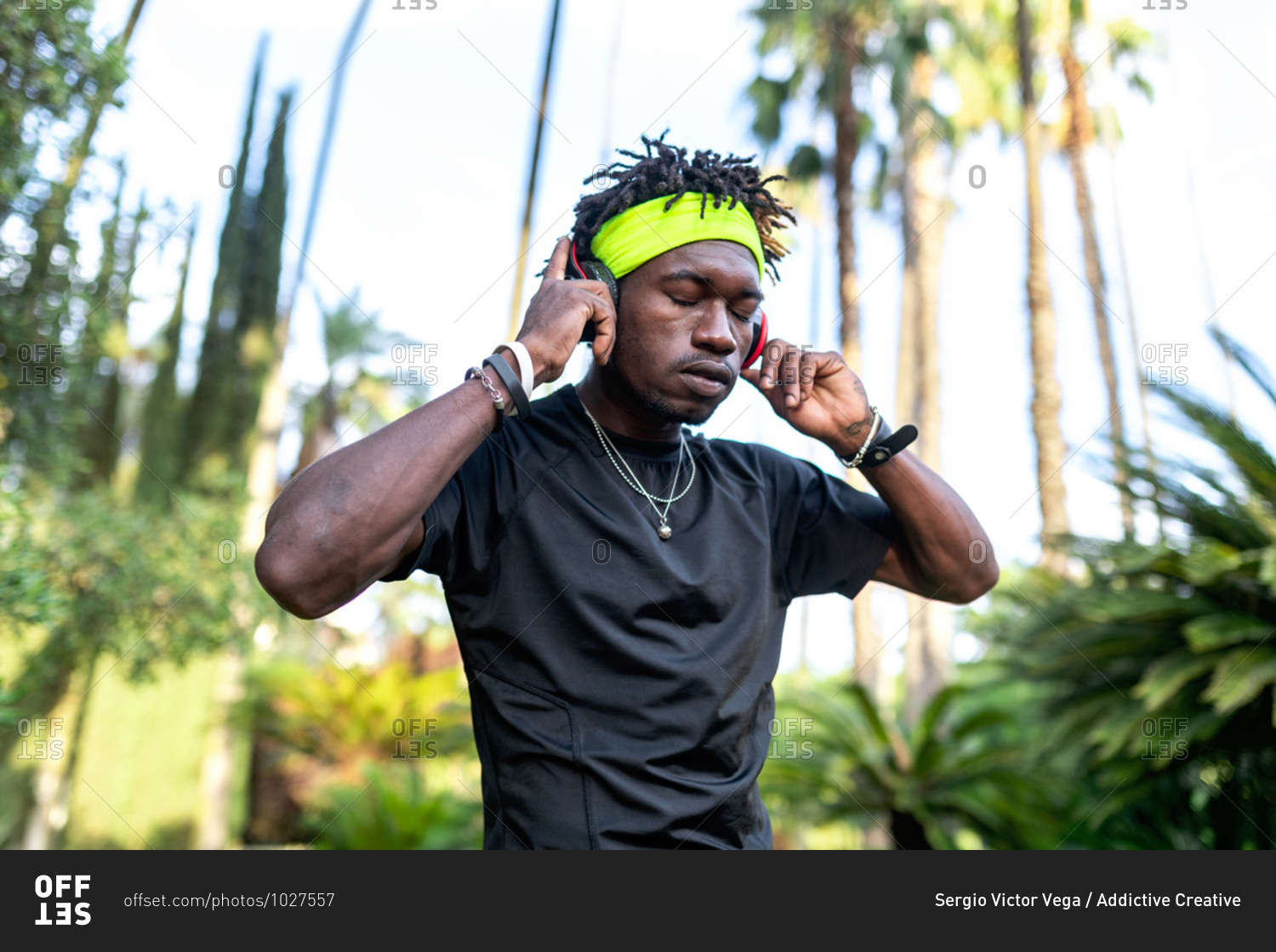 Young African American male athlete in sportswear listening to music with his headphones keeping hands on the headphones and closed eyes while standing in green tropical park