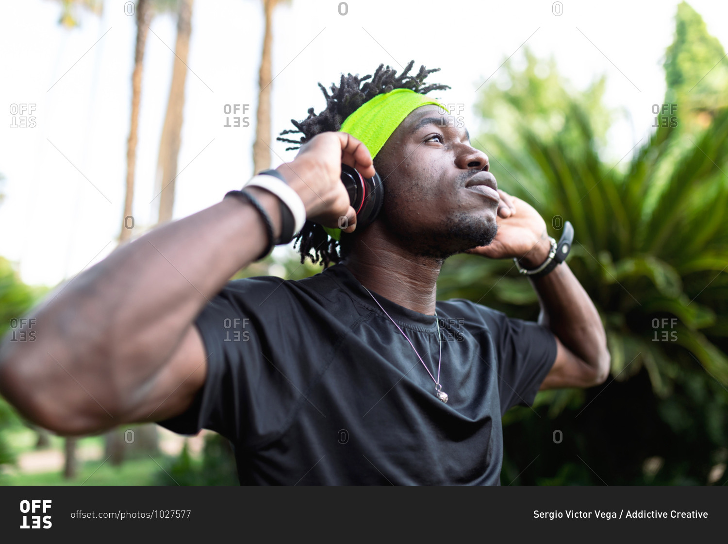 Young African American male athlete in sportswear listening to music with his headphones keeping hands on the headphones and looking away while standing in green tropical park