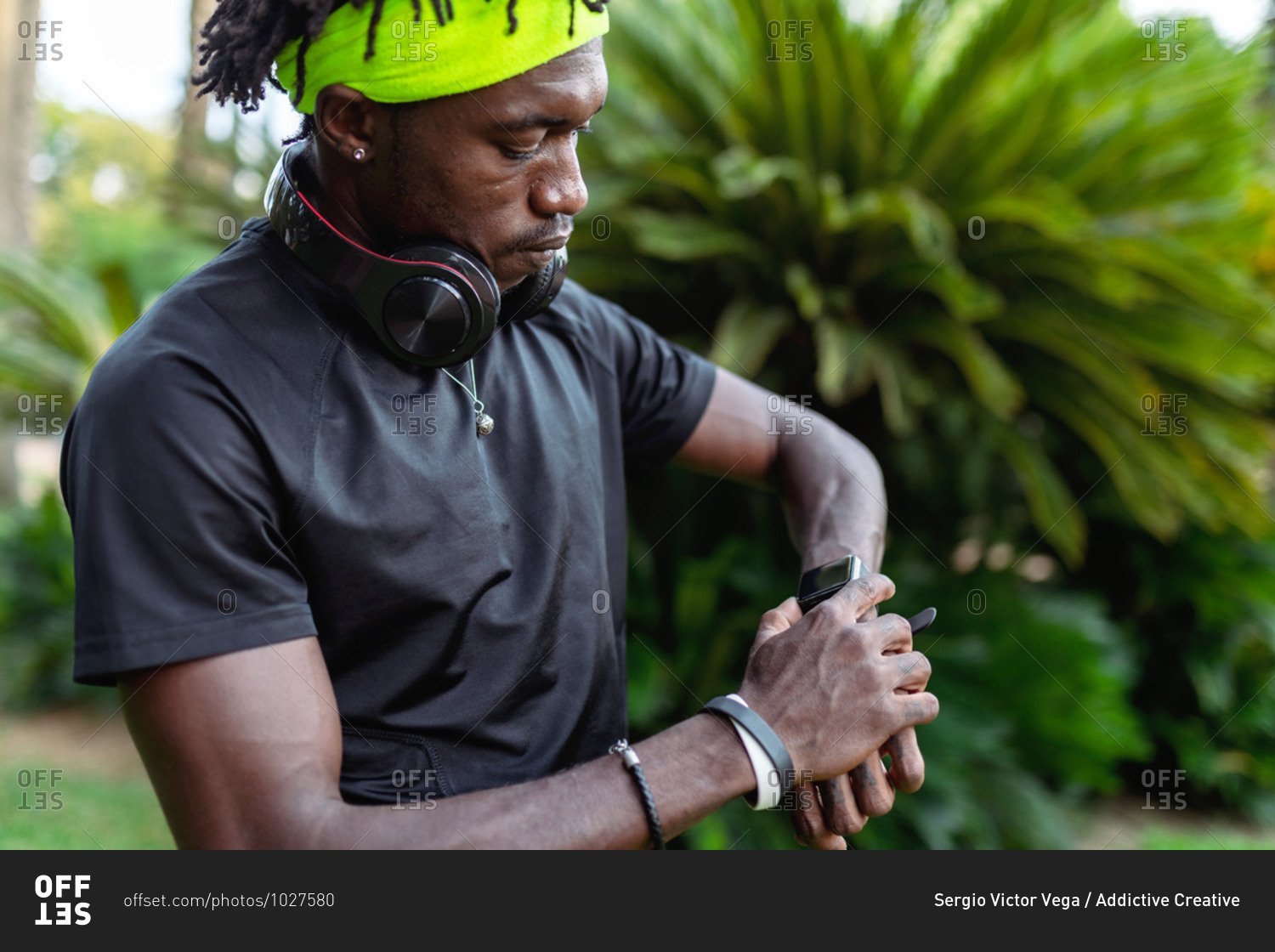 Stylish young African American male runner with headphones on neck checking sport results on fitness tracker during training in green tropical park