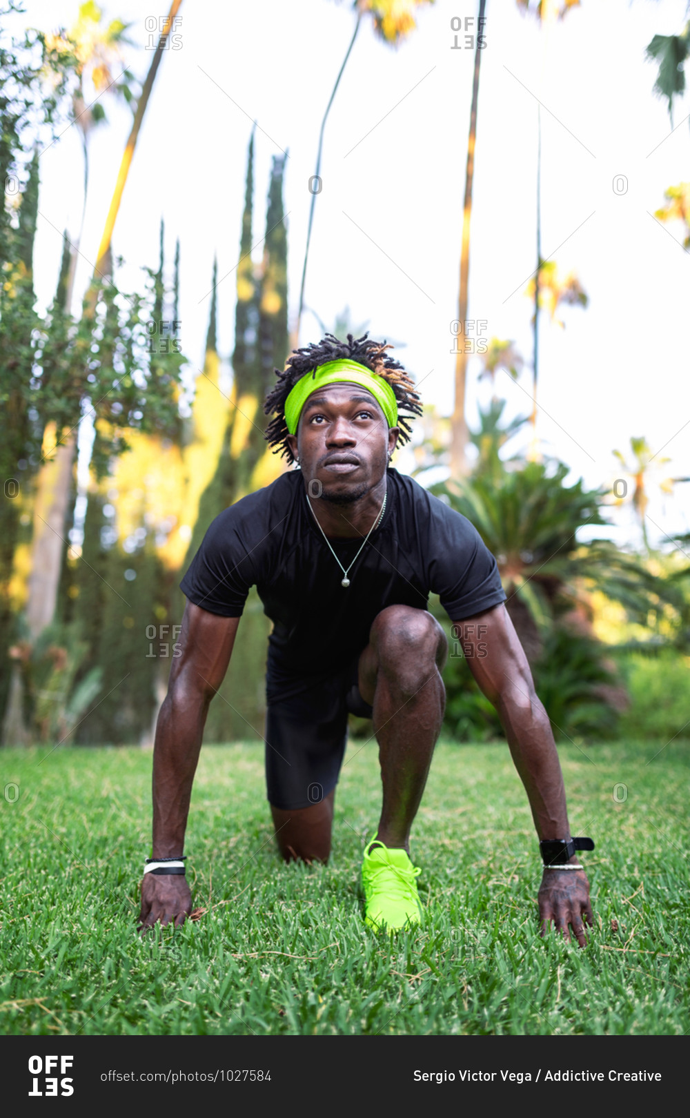 Determined young African American male athlete in stylish sportswear standing in low start position on green lawn while preparing for running in park