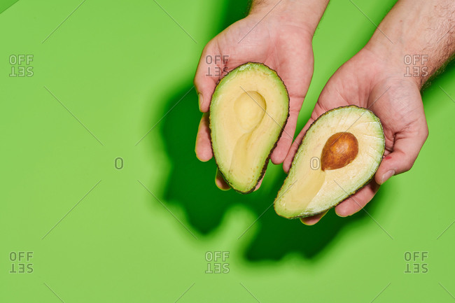 From above of crop unrecognizable person holding halves of fresh ripe green avocado with seed against bright green background