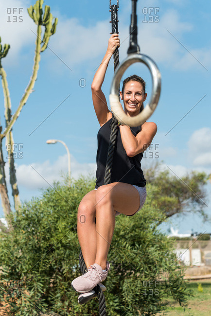 Muscular female athlete climbing rope while training on sports ground in city and looking at camera