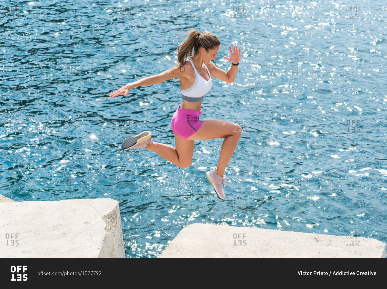 Side view of strong female athlete in sportswear in moment of jumping on breakwater boulders during training at seaside in summer
