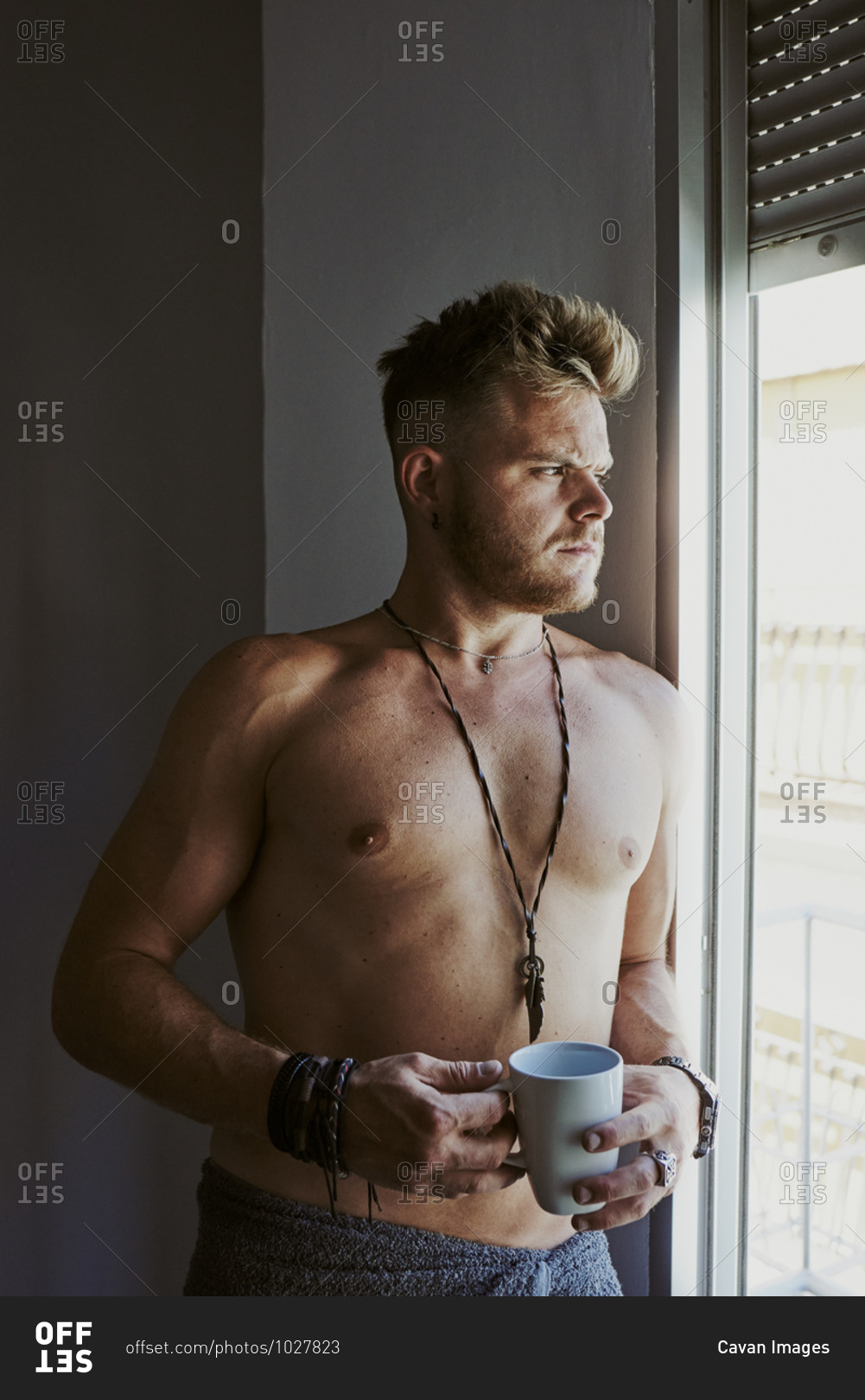 a blond man with no shirt on takes a cup of coffee while looking out the window