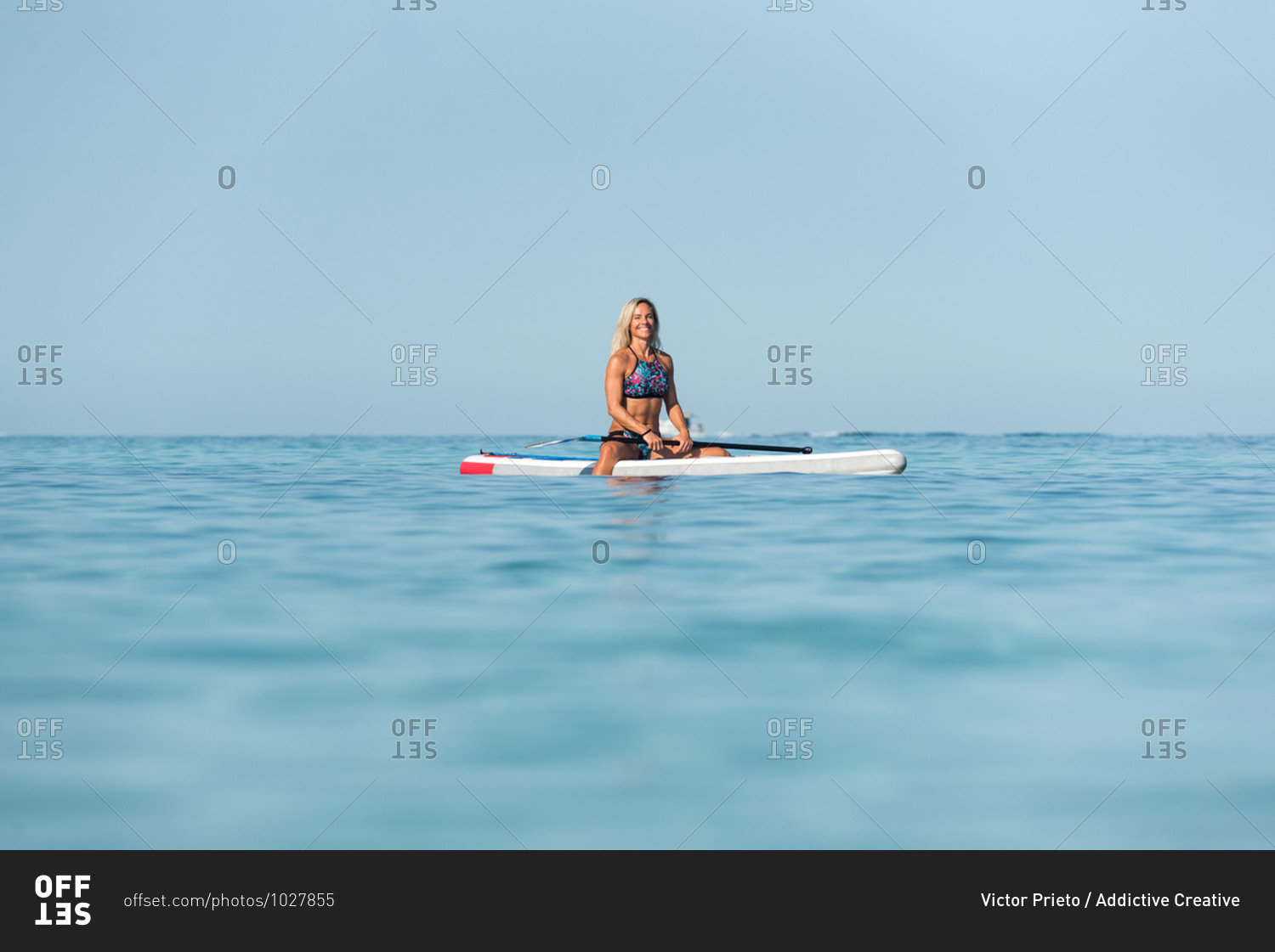Fit happy female surfer sitting on surfboard with paddle and relaxing during training in calm sea in summer looking at camera