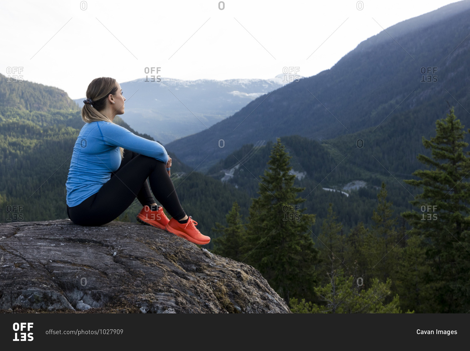 Strong sportswoman sitting and taking a break from trail running against forested mountain ridge during fitness workout in countryside