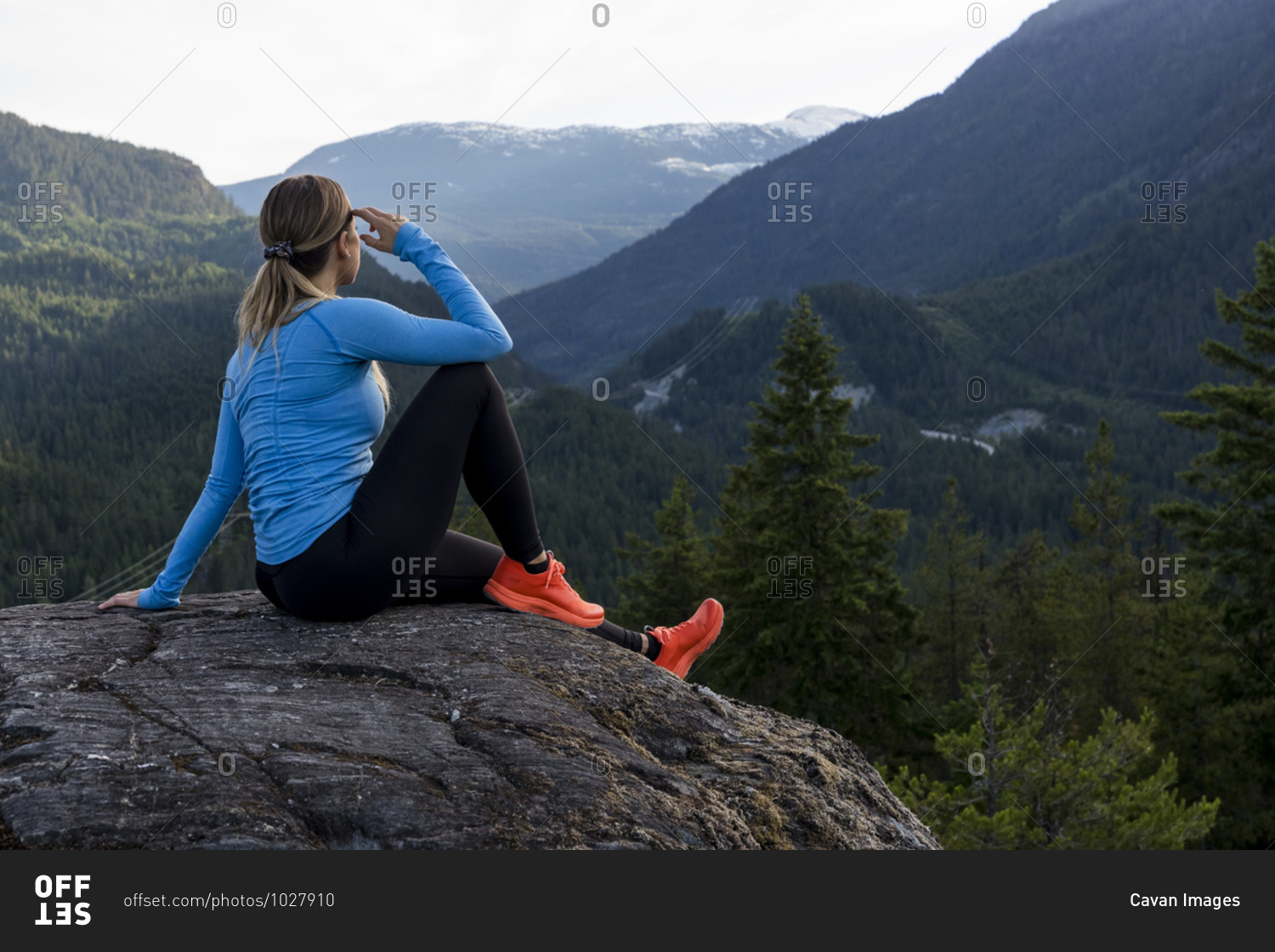 Female athlete sitting and taking a break from trail running against forested mountain ridge during fitness workout in countryside