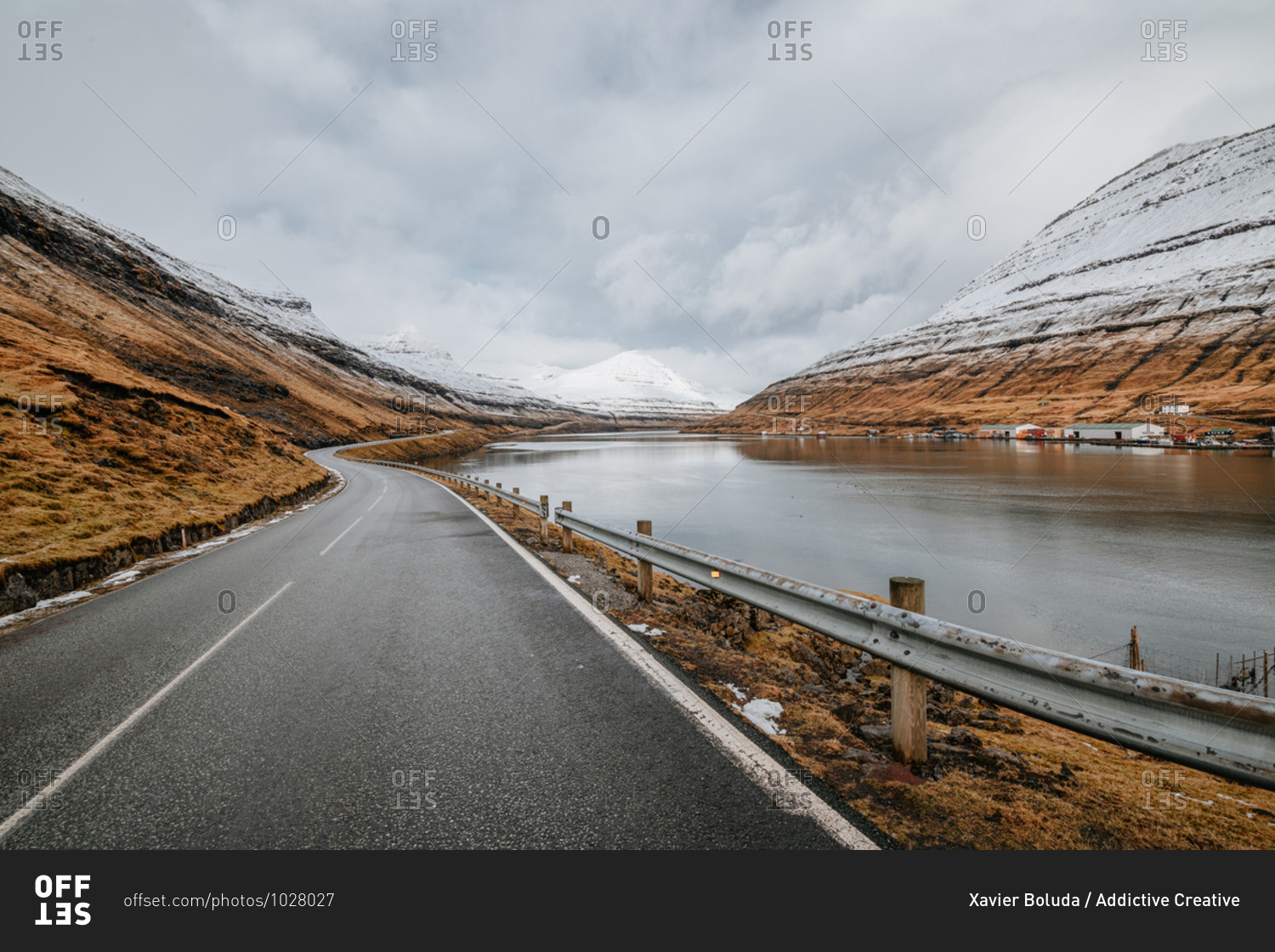 Spectacular view of wet winding road located near river and mountain slope with dry grass in winter on Faroe Islands