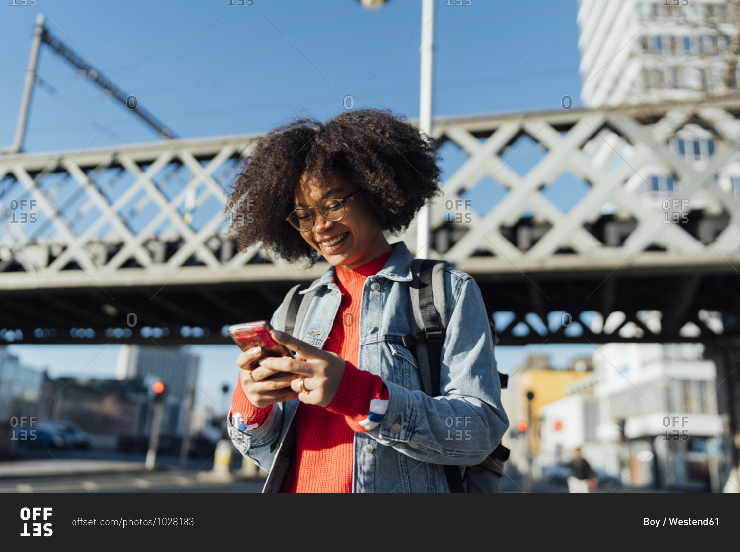 Smiling afro young woman using smart phone while standing against bridge in city