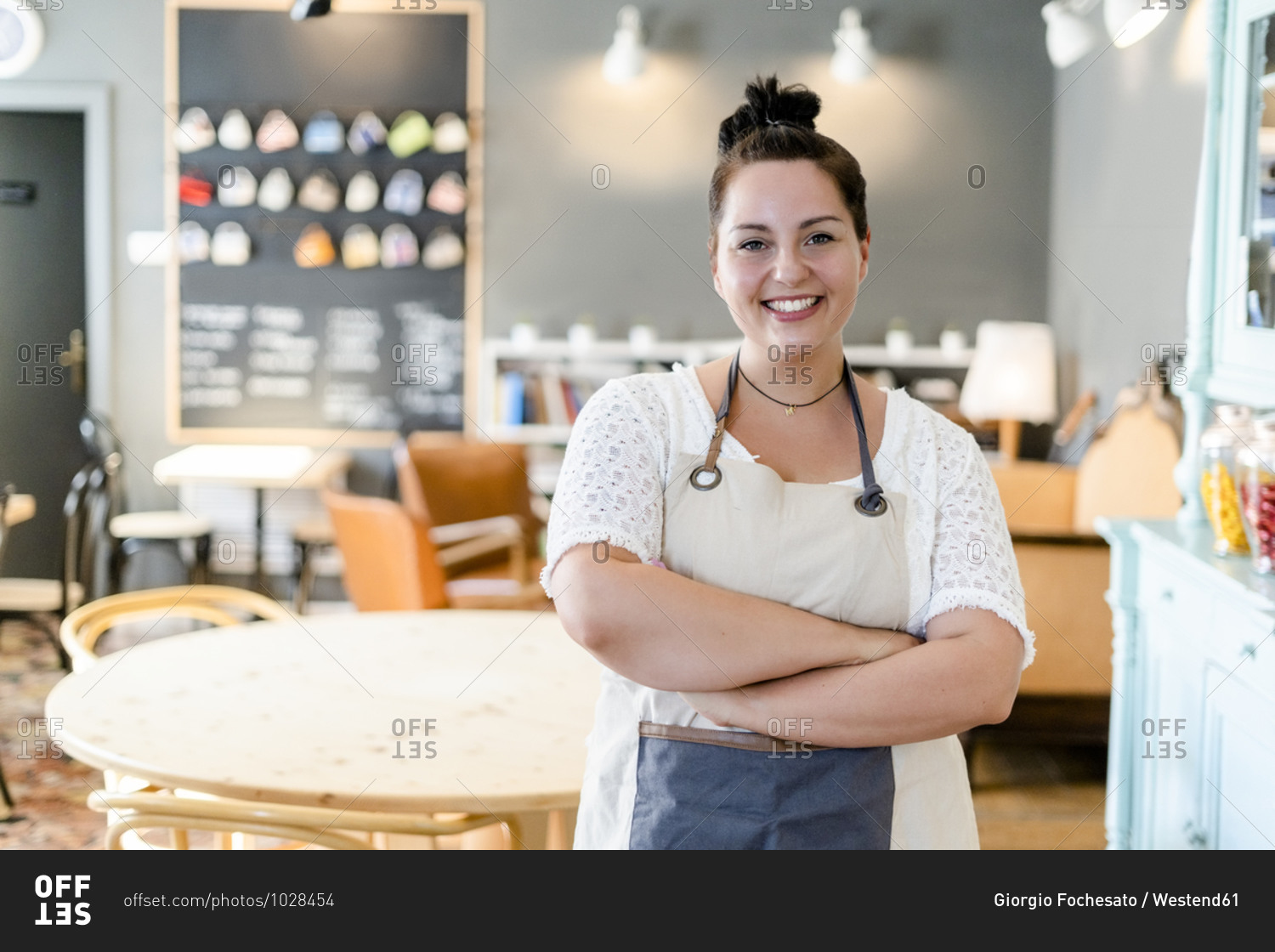 Smiling female owner with arms crossed standing against furniture in coffee shop