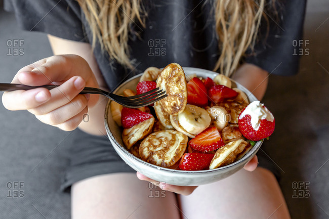 Mid section of teenage girl eating bowl of mini pancakes with strawberries and bananas