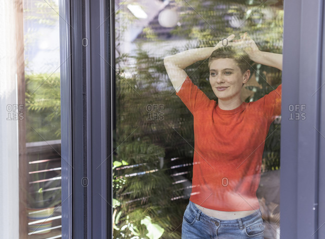 Thoughtful woman with arms raised looking through window while standing at home seen through glass