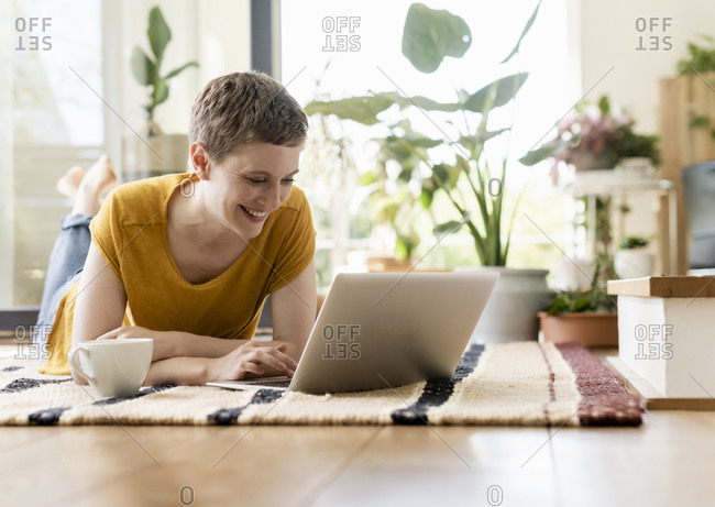 Smiling mid adult woman with short hair using laptop while relaxing on carpet at home