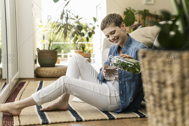 Smiling woman with short hair pouring drink in glass while sitting on carpet at home