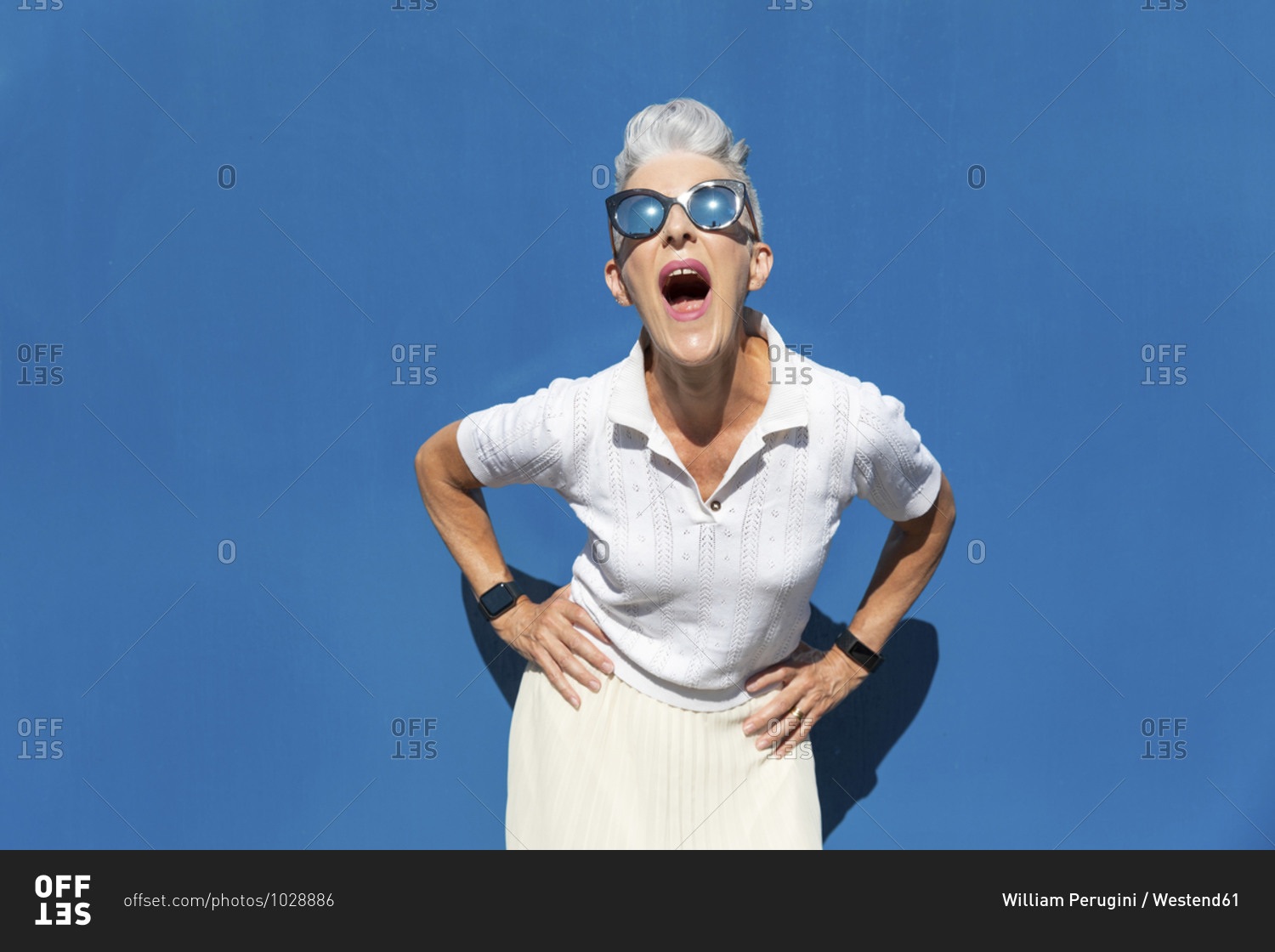 Woman with mouth open and hand on hip standing against blue
wall during sunny day stock photo - OFFSET