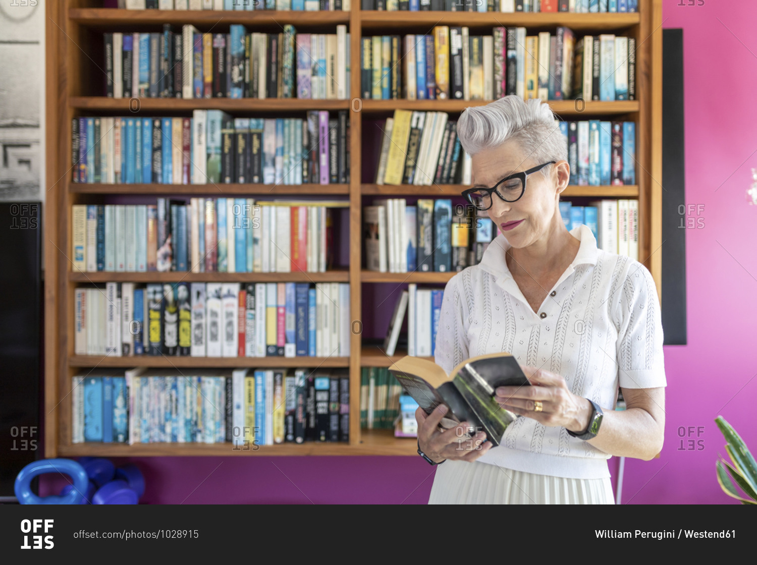 Active senior woman reading book while standing against bookshelf at home
