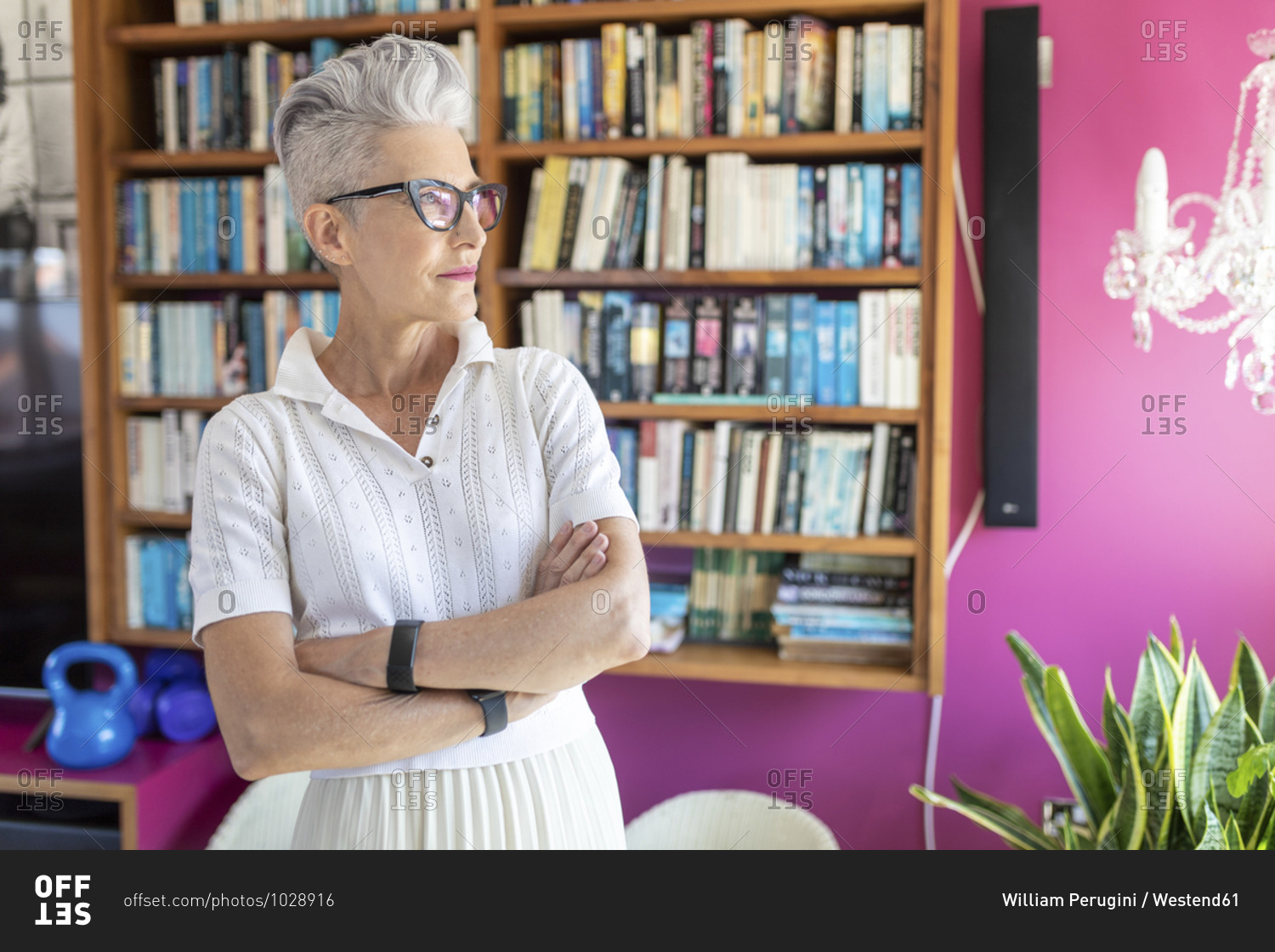 Senior female with arms crossed standing against bookshelf at home