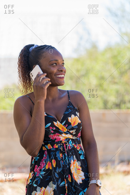 Portrait of happy afro American woman with a ponytail and braided hair wearing colorful dress talking on the mobile phone outdoors