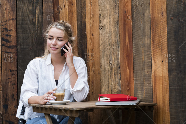 Young blond woman sitting alone in a cafe, using mobile phone, working remotely.