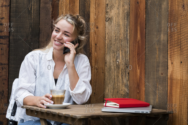 Young blond woman sitting alone in a cafe, using mobile phone, working remotely.