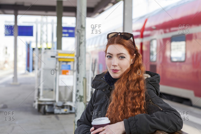 Thoughtful young woman with long red hair wears a coat and holds a coffee cup while waiting on a bench at a train station