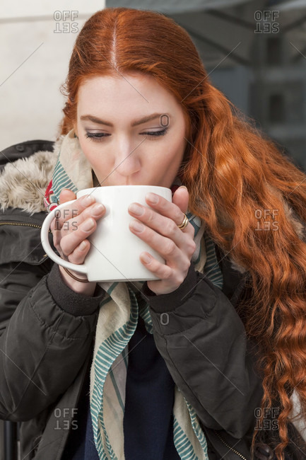 Happy red haired woman holding large white mug while seated at table outside in cool weather clothing