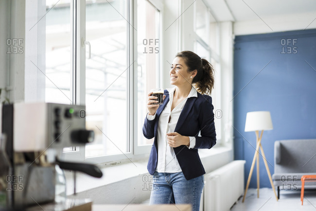 Happy businesswoman with glass of coffee in a loft looking out of window