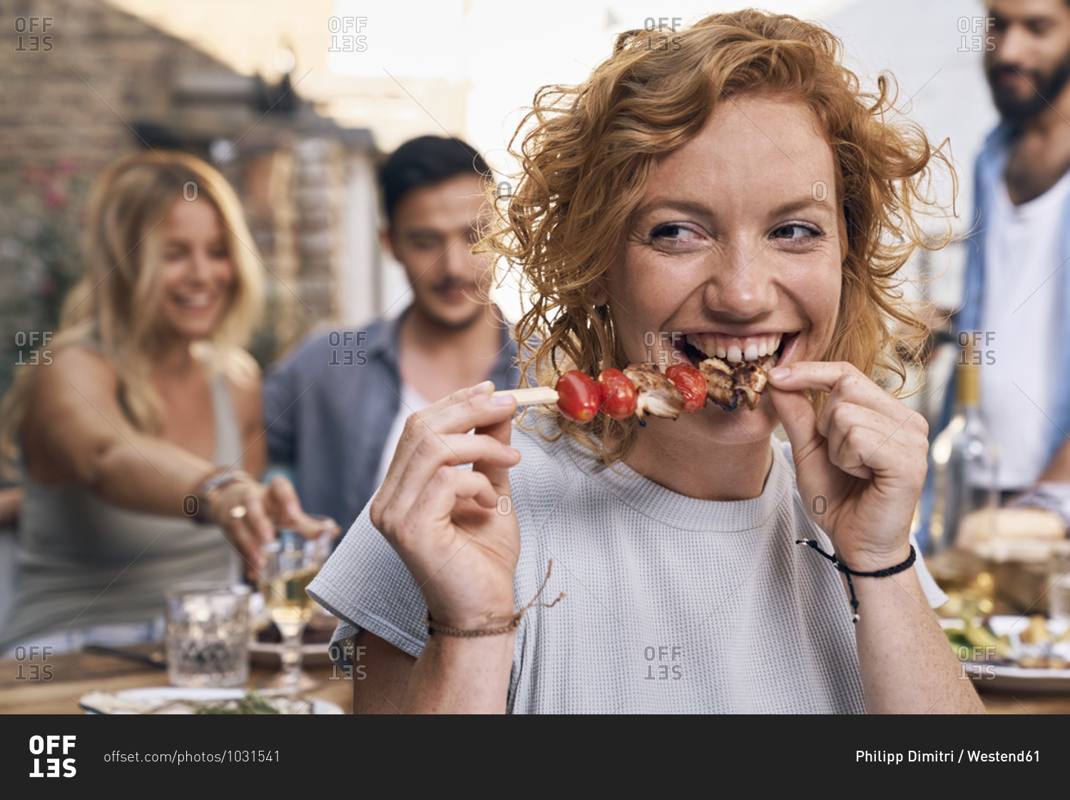 Young woman eating meat skewer at a backyard party with friends