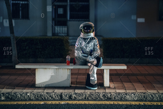 Spaceman sitting on bench at a bus stop at night with soft drink