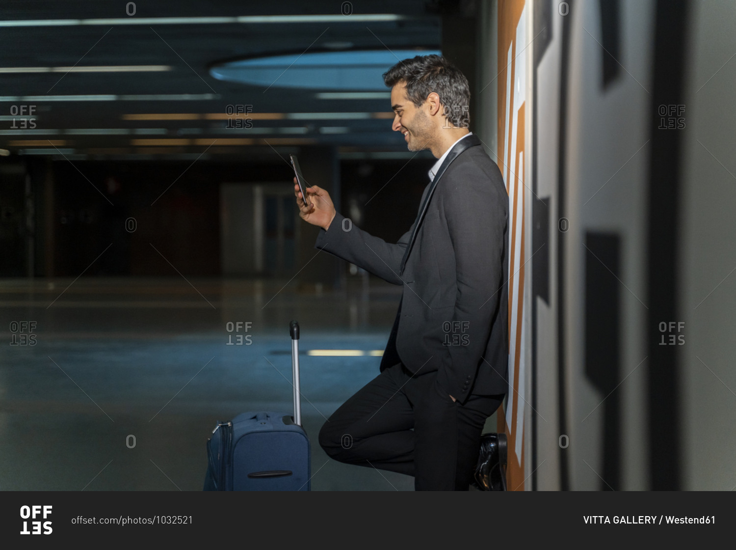 Smiling male entrepreneur using mobile phone while standing with suitcase by wall at station