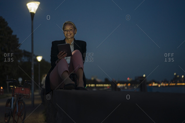 Smiling beautiful female entrepreneur using digital tablet while sitting on retaining wall against sky at night