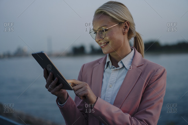 Smiling beautiful blond businesswoman using digital tablet against river at dusk