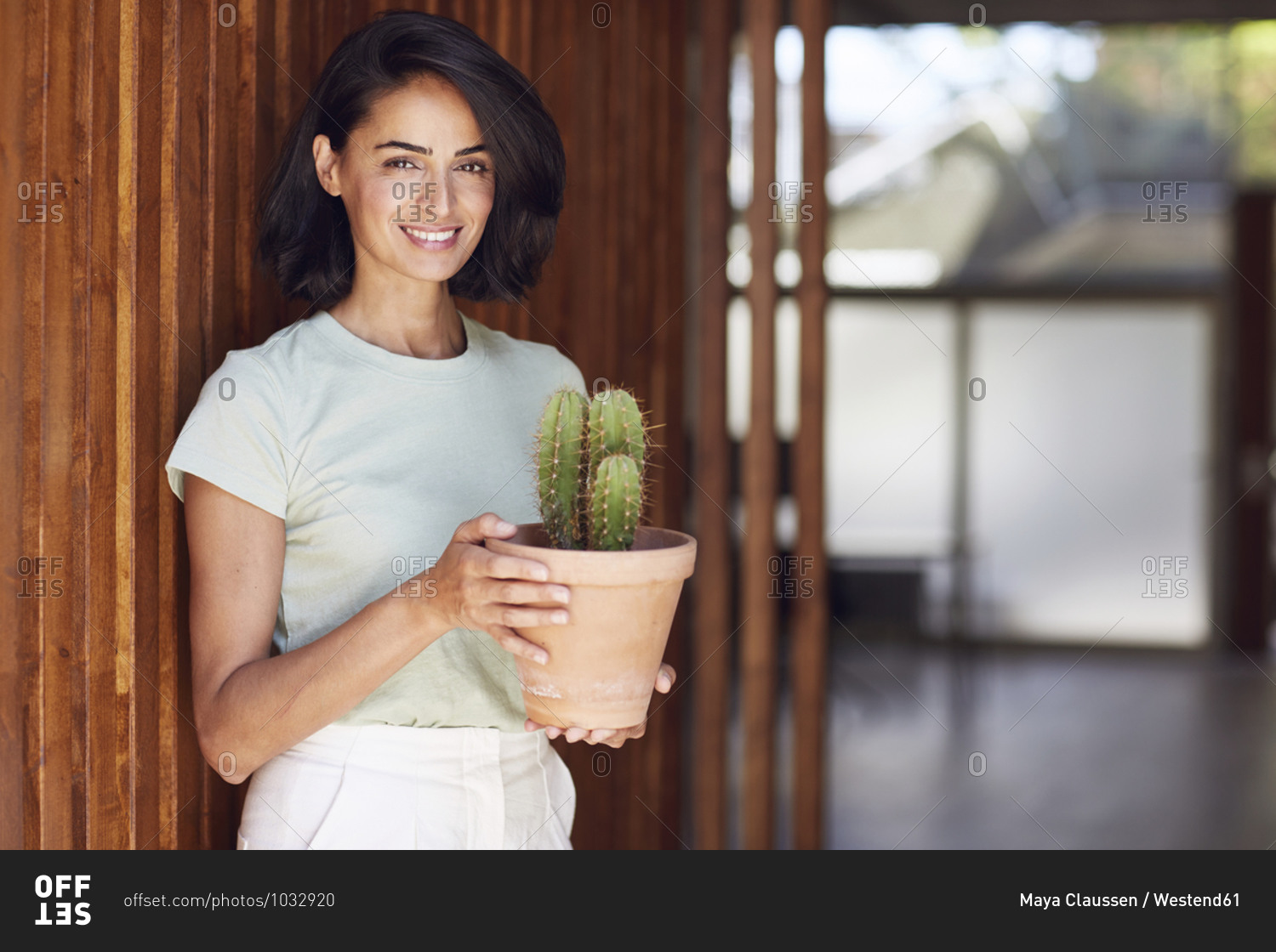 Smiling female entrepreneur holding cactus plant while standing by wooden wall in office