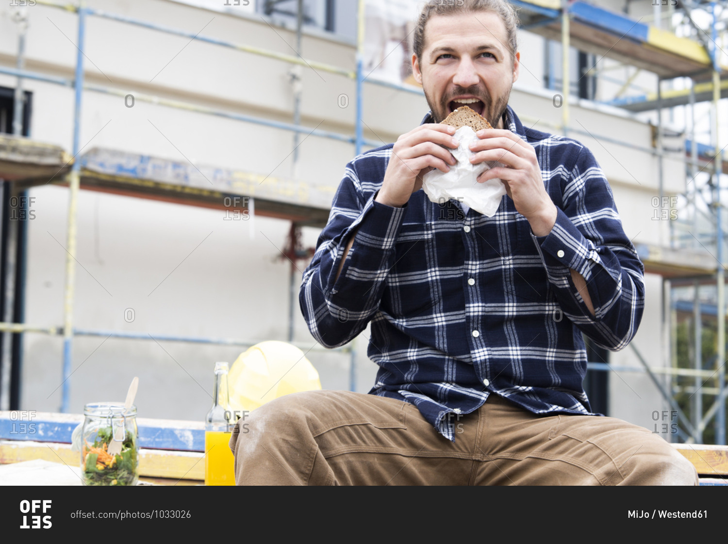 Construction worker eating bread while sitting against building at construction site