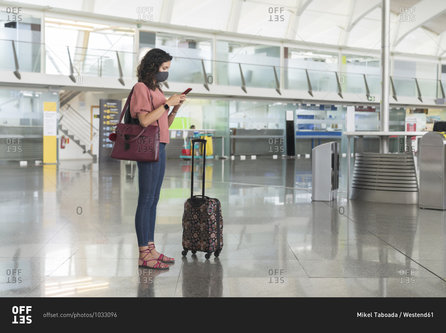 Young woman with protective face mask text messaging on smart phone while standing at airport