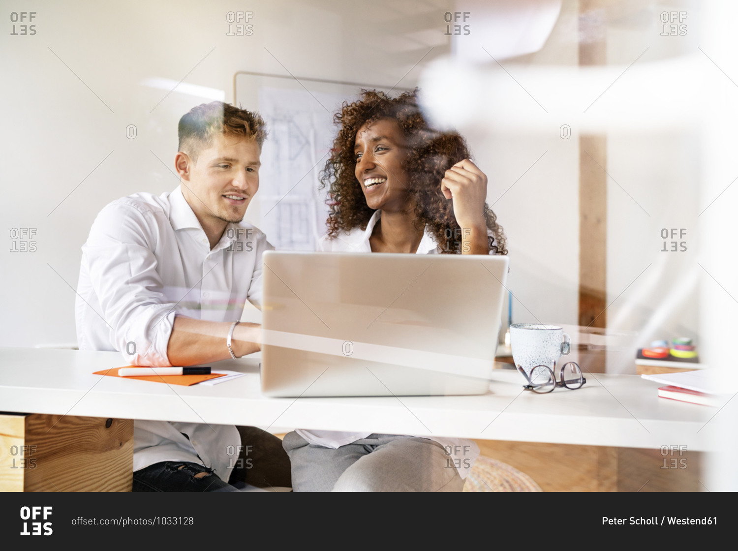 Smiling colleagues discussing over laptop on desk in office seen through window