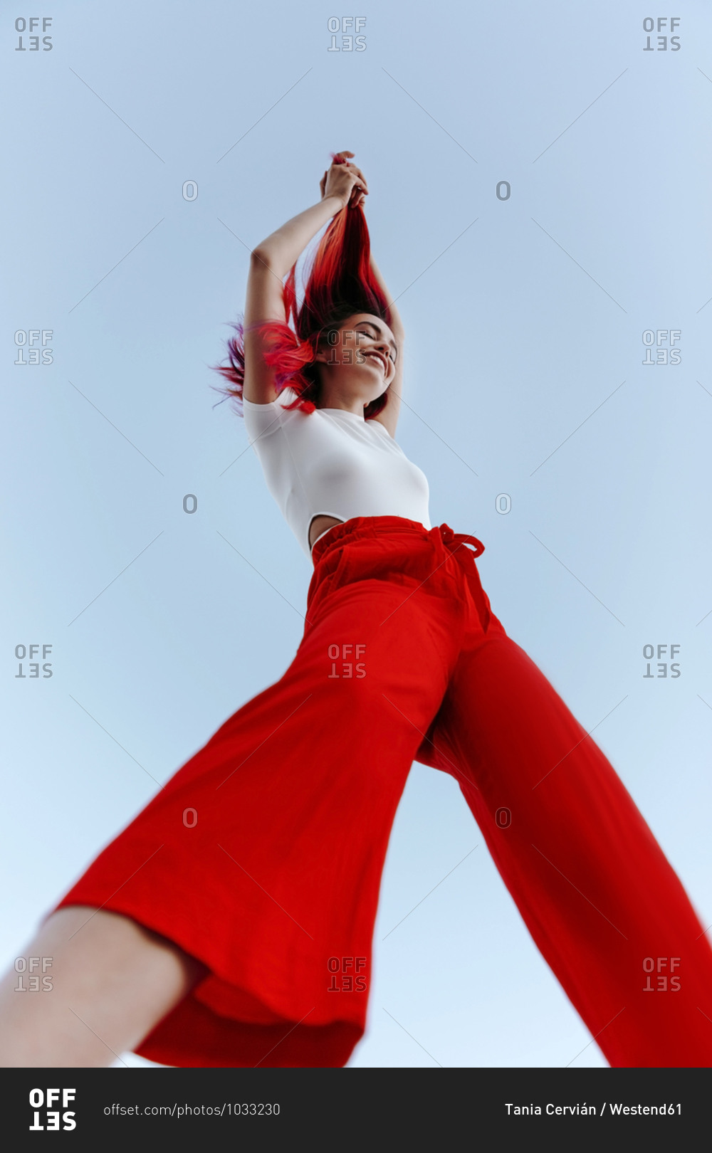 Smiling young woman pulling dyed red hair up while standing against clear sky