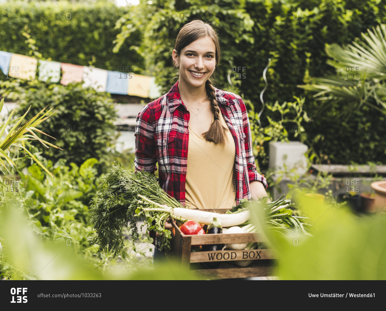Smiling woman carrying vegetables in crate while standing against plants at garden