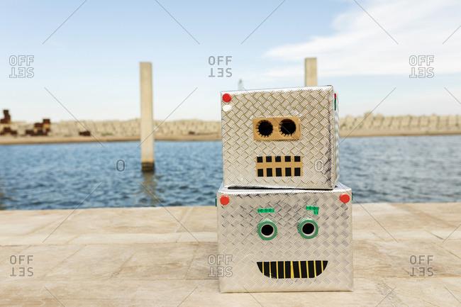 Robot masks made of boxes on footpath against water