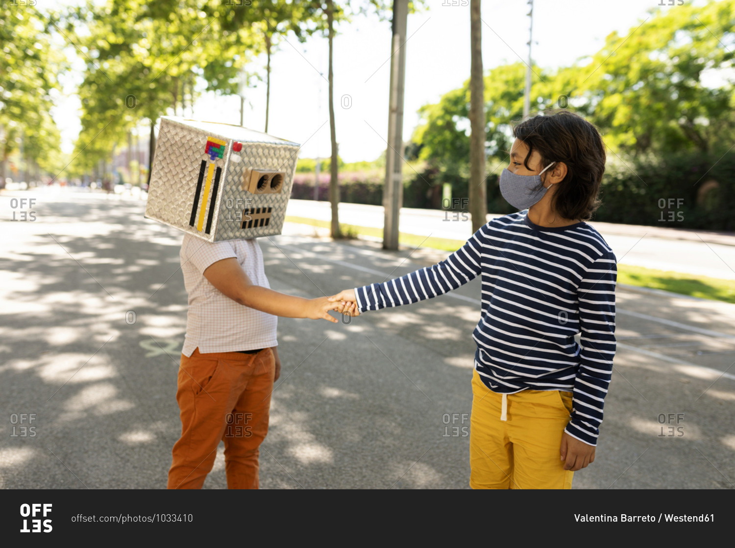 Boy in face mask giving handshake to friend wearing robot costume while standing on road
