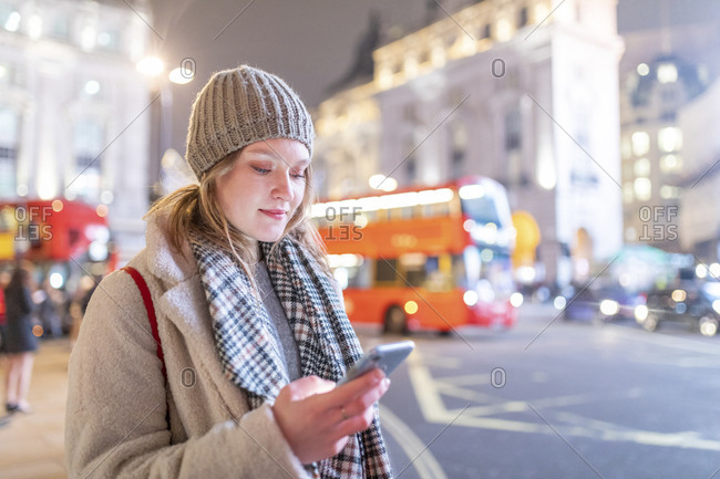 Young woman using smart phone while standing by street in Piccadilly Circus at night