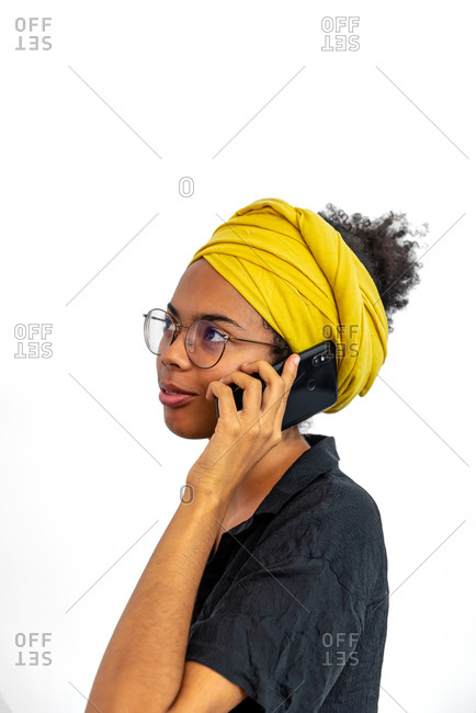 Portrait of young woman wearing glasses and yellow head scarf and speaking on mobile phone over white background
