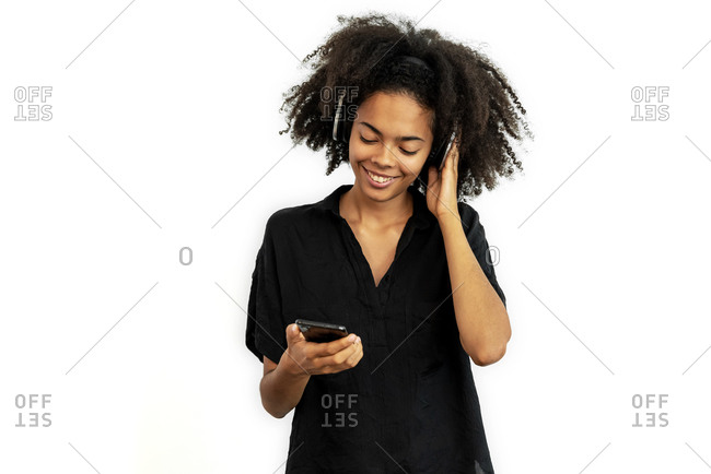 Young mixed race woman with headphones listening music on a phone while posing against white blank wall