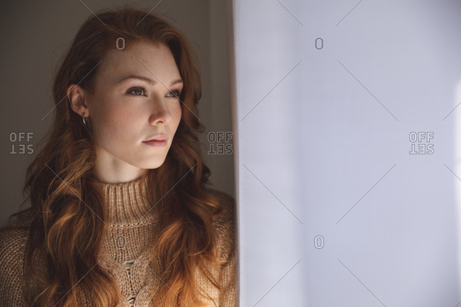 An attractive, ginger Caucasian woman spending time at home, in living room, looking out of the window. Social distancing during Covid 19 Coronavirus quarantine lockdown.