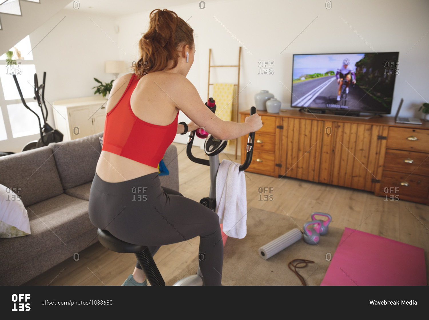Caucasian woman spending time at home, in living room, exercising on stationary bike, watching tv. Social distancing during Covid 19 Coronavirus quarantine lockdown.