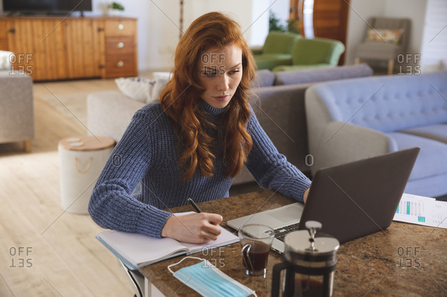 Caucasian woman spending time at home, in the kitchen, working from home, using her laptop and writing. Social distancing during Covid 19 Coronavirus quarantine lockdown.