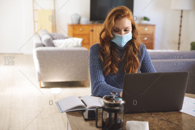 Caucasian woman spending time at home, in the kitchen, working from home, using her laptop,  wearing a face mask. Social distancing during Covid 19 Coronavirus quarantine lockdown.
