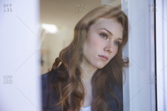 An attractive, ginger Caucasian woman spending time at home, in living room, looking out of the window. Social distancing during Covid 19 Coronavirus quarantine lockdown.