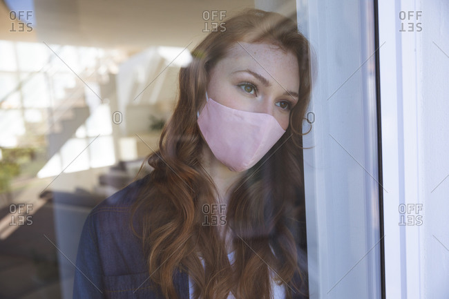 An attractive, ginger Caucasian woman spending time at home, in living room, looking out of the window, wearing a face mask. Social distancing during Covid 19 Coronavirus quarantine lockdown.
