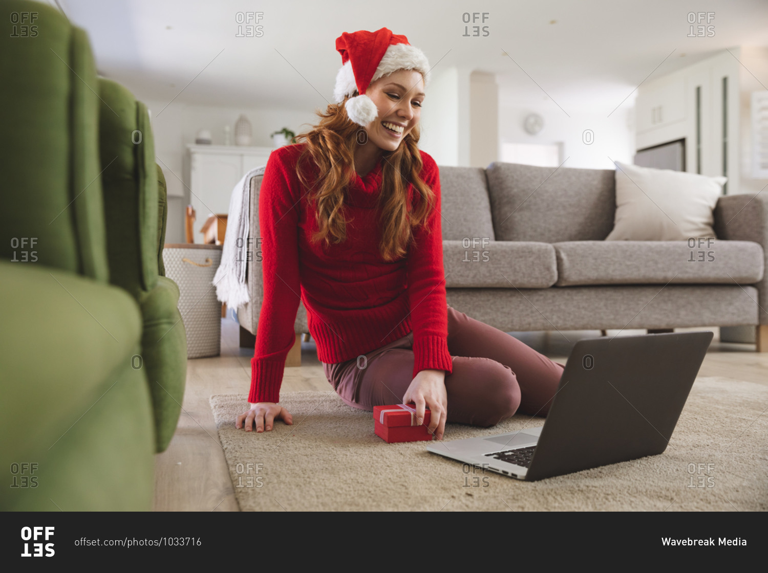 Caucasian woman spending time at home, in living room, smiling, wearing Christmas hat, holding a gift during a video call. Social distancing during Covid 19 Coronavirus quarantine lockdown.